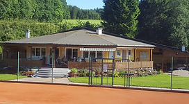 Tennisplatz Schneegattern in Lengau: Ein charmantes Holzblockhaus mit Veranda, umgeben von üppigem Grün. Das Gebäude befindet sich inmitten einer idyllischen Landschaft mit Wiesen und Wäldern.