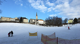 Skilift Maria Schmolln in Maria Schmolln: Eine verschneite Landschaft mit einer Kirche oder einem Kirchturm im Vordergrund. Im Hintergrund sind Gebäude zu sehen, die auf einen Ort hinweisen. Im Vordergrund befinden sich Menschen, die vermutlich auf der Eisfläche Schlittschuhlaufen.