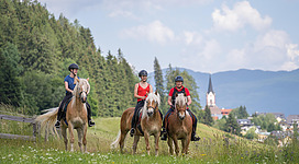 Drei Reiterinnen auf Pferden genießen die Natur. Im Hintergrund sind grüne Wiesen und ein Dorf mit einer Kirche zu sehen.