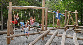 Ein Spielplatz aus Holz mit mehreren Kindern, die spielen und schwingen. Die Umgebung ist begrünt und naturnah.