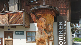 Ein traditionelles Gebäude mit einer Holzkulptur von einem Pferd an der Wand. Vor dem Eingang befinden sich bunte Blumenbeete und grüne Sträucher.