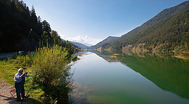 Ein schöner See mit ruhigem, klarem Wasser und umgeben von Bergen. Eine Person fischt am Ufer in der Sonne.
