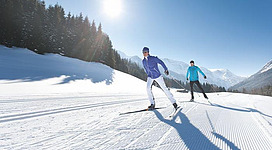 Zwei Männer skaten auf einer schneebedeckten Piste bei sonnigem Wetter. Im Hintergrund sind verschneite Berge und Nadelbäume zu sehen.