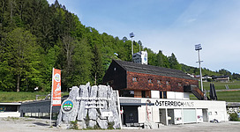 Ein modernes Gebäude im alpinen Stil, umgeben von Bäumen und einer Sportanlage. Der Schriftzug „Österreich Haus“ ist gut sichtbar.