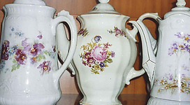 Drei elegante Porzellan-Kännchen mit Blumenmotiven stehen nebeneinander. Die Kännchen haben verschiedene Formen und detaillierte Verzierungen.