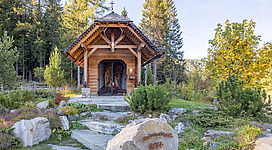 Eine kleine Holzkirche umgeben von Bäumen und buntem Laub. Der Eingang ist architektonisch ansprechend und die Landschaft wirkt einladend.
