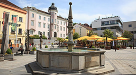 Ein schöner Platz mit einem Brunnen in der Mitte. Umgeben von modernen und historischen Gebäuden sowie gemütlichen Sitzbereichen.
