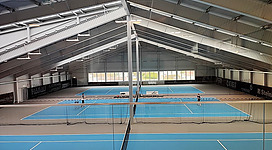 Tennis-Zentrum Danubis in Mauthausen: Eine große, überdachte Sportanlage mit mehreren Tennisplätzen. Im Inneren gibt es ein geräumiges Hallengebäude mit hoher Decke, das die Tennisplätze beherbergt. Die Böden der Plätze sind in blau- und hellgrauen Farbtönen gehalten. Insgesamt bietet die Anlage Möglichkeiten für sportliche Betätigung in einer modernen und zweckmäßigen Umgebung.