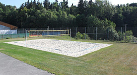 Beach-Volleyball in St. Martin im Mühlkreis: Ein Sportfeld mit Sandplatz und Volleyballnetz. Das Feld ist von einem Maschendrahtzaun umgeben und liegt inmitten einer waldigen Umgebung mit Bäumen. Der Rasen rund um den Platz ist sorgfältig gepflegt.