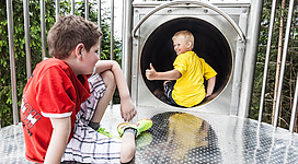 Zwei Kinder spielen auf einem Spielplatz. Ein Junge rutscht aus einem Tunnel, während der andere entspannt daneben sitzt.
