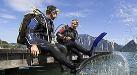 Tauch- & Paddlestation Wolfgangsee in St. Wolfgang im Salzkammergut: Das Bild zeigt zwei Menschen in Tauchtausrüstung, die auf einem Holzsteg am Wasser sitzen. Sie scheinen eine Pause von einer Tauchaktivität zu machen und genießen die schöne Umgebung mit Bergrücken und Himmel im Hintergrund.