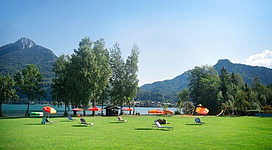 Eine entspannende Wiese am Wasser mit Liegestühlen und Bäumen. Im Hintergrund sind Berge und ein klarer blauer Himmel sichtbar.