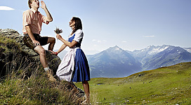 Ein Junge sitzt auf einem Felsen und schaut zur Seite. Ein Mädchen in traditioneller Tracht reicht ihm Blumen vor einer malerischen Berglandschaft.