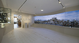 Stille Nacht Krippe in Ried im Innkreis: Ein großer, heller Raum mit einer Wand, die eine detaillierte, panoramische Landschaftsdarstellung zeigt. Der Boden ist mit weißen Fliesen ausgelegt, und die Decke ist mit Scheinwerfern ausgestattet. Einige Gestaltungselemente und Möbel sind im Vordergrund zu sehen.