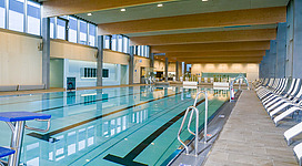 Freizeitbad Ried in Ried im Innkreis: Großer Innenraum mit hoher Decke, der als Schwimmhalle dient. Das Becken ist groß und zentral platziert, umgeben von Sitzgelegenheiten und Treppen. Die Beleuchtung ist warm und schafft eine einladende Atmosphäre. Die Architektur wirkt modern und funktional.