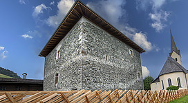 Ein historisches Gebäude aus Stein mit einem Holzdach. Im Hintergrund ist ein Kirchturm zu sehen unter einem teilweise bewölkten Himmel.