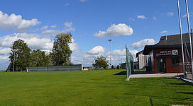 Faustballplatz in Höhnhart: Eine große grüne Rasenfläche, umgeben von Bäumen und bewölktem Himmel. Auf einer Seite befindet sich ein Gebäude mit einer Beschriftung.