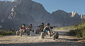 Eine Gruppe von Kindern fährt mit großen Karts auf einem Kiesweg. Im Hintergrund sind beeindruckende Berge und ein klarer Himmel zu sehen.