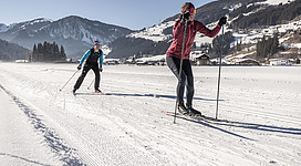 Zwei Langläuferinnen gleiten über eine schneebedeckte Fläche in den Bergen. Die klare, sonnige Winterlandschaft umgibt sie.