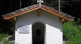 Eine kleine, weiße Kapelle mit einem Dach aus Holz. Sie befindet sich umgeben von Bäumen und Gras in der Natur.