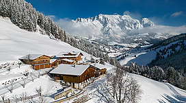 Eine malerische Winterlandschaft mit schneebedeckten Hügeln und traditionellen Holzhäusern. Im Hintergrund ragen majestätische Berge unter einem klaren blauen Himmel hervor.