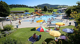Ein sonniger Freizeitpark mit einem großen Pool und Rutschen. Viele Menschen entspannen sich auf Liegen und genießen die Zeit im Freien.