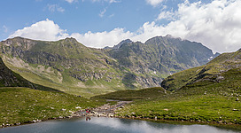 Eine friedliche Berglandschaft mit einem klaren See im Vordergrund. Die umliegenden Berge sind grün und majestätisch, unter einem blauen Himmel mit wenigen Wolken.