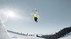 Ein Skifahrer führt einen spektakulären Sprung in der Luft aus. Die Landschaft ist verschneit mit Bergen im Hintergrund und einem klaren Himmel.