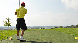 Golfclub Perg-Karlingberg in Perg: Eine männliche Person in einer gelben Jacke steht auf einem Golfplatz. Im Hintergrund ist eine grüne Landschaft und Berge zu sehen.