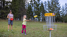 Ein Kind spielt Disc Golf und wirft eine Scheibe in Richtung Korb. Eine erwachsene Begleitperson steht im Hintergrund und beobachtet das Spiel.