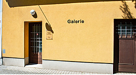 Galerie im Zeughaus in Perg: An gelb gestrichene Wand mit dem Schriftzug "Galerie" darauf. Davor steht eine Eingangstür, die von einem kleinen, geschlossenen Vorbau überdacht ist. Oberhalb des Eingangs ist eine Kugel-Außenbeleuchtung angebracht. Der Boden vor dem Eingang ist mit Pflastersteinen ausgelegt.