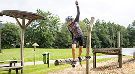 Fitness- und Motorikpark Schneegattern in Lengau: Eine Person auf einem Fahrrad, die sich über einen Holzsteg bewegt, der von einem Grünraum umgeben ist. Hinter ihr befinden sich weitere Holzkonstruktionen und eine grüne Umgebung mit Bäumen.