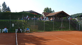 Tennisplatz Eggelsberg in Eggelsberg: Ein Tennisplatz mit grünen Umzäunungen, an dem sich mehrere Personen in Sportbekleidung befinden. Einige sitzen auf Bänken und beobachten das Geschehen, während andere auf dem Platz spielen.