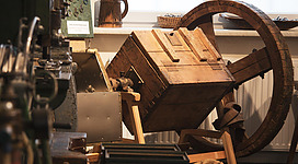 Eine alte Holzkiste steht in einem Raum voller Maschinen. Daneben ist ein großes Rad sichtbar, das Teil der Ausrüstung ist.