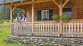 Ein freundliches Paar steht auf der Veranda eines Holzhauses. Der Garten ist grün und gepflegt, umgeben von Bergen.