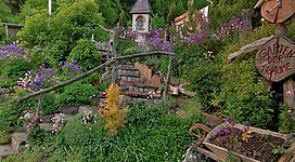 Ein wunderschöner Garten mit bunten Blumen und üppigem Grün. Im Hintergrund steht ein kleiner Pavillon und ein Schild mit der Aufschrift „Garten der Träume“.
