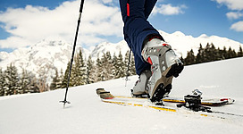 Nahaufnahme von einer Person beim Skitourengehen: Ein Bein in einer Skitourenbindung in Bewegung, Skistöcke und verschneite Winterlandschaft mit Bergen und Tannen im Hintergrund.