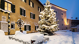 Ein verschneiter Innenhof mit einem geschmückten Weihnachtsbaum und alten Gebäuden. Die Szene strahlt eine ruhige Winteratmosphäre aus.