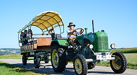 Ein Mann in Lederhose und Hut fährt einen Oldtimer Traktor mit Anhänger durch eine grüne Landschaft. Der Anhänger ist mit einer Plane überdacht und bietet Sitzgelegenheiten, einige Personen fahren mit.