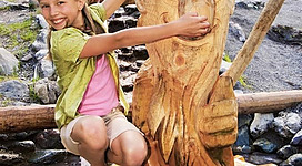 Ein Mädchen spielt fröhlich auf einem Holzspielgerät in der Natur. Im Hintergrund sind andere Kinder und eine schöne Berglandschaft zu sehen.