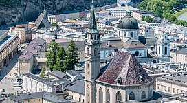 Eine malerische Stadtansicht mit historischer Architektur und grünen Bäumen. Im Vordergrund steht eine markante Kirche mit spitzem Turm.