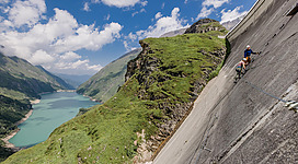 Ein Kletterer erklimmt eine steile Felswand mit Blick auf einen glitzernden Stausee und die umliegenden Berge. Der Himmel ist blau mit einigen Wolken.