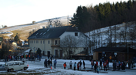 Eisstockbahnen in Lembach im Mühlkreis: Eine historische Gebäudeanlage mit Holz- und Steinbauten, umgeben von Bäumen und Vegetation. Vor dem Gebäudeeingang stehen mehrere Personengruppen, die vermutlich auf Aktivitäten oder Veranstaltungen in diesem Ort warten. Die Landschaft im Hintergrund ist von schneebedeckten Hügeln und Wäldern geprägt.
