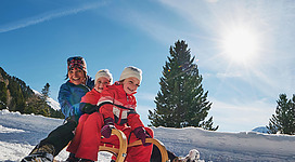Eine fröhliche Familie fährt mit einem Schlitten den verschneiten Hang hinunter. Die Sonne scheint und der Himmel ist blau.