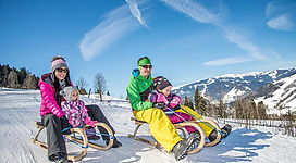 Familie mit zwei Kindern beim Rodeln im Schnee an einem sonnigen Wintertag in den Bergen.