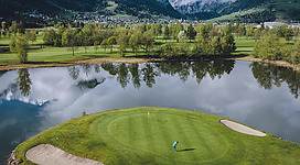 Eine grüne Golfanlage mit einem kleinen See und Bergen im Hintergrund. Der Platz ist umgeben von Bäumen und reflektiert im Wasser.