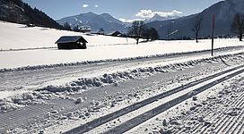Eine verschneite Landschaft mit einem kleinen Holzhäuschen. Im Hintergrund sind Berge und ein klarer blauer Himmel zu sehen.