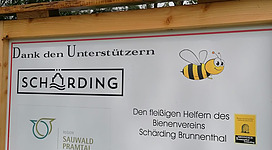 Bienenlehrpfad in Schärding: Eine Außenanzeige mit Informationen über einen Bienenverein. Im Vordergrund ist ein Holzrahmen zu sehen, auf dem der Name "Scharding" und ein Bienenlogo abgebildet sind. Außerdem sind weitere Details zu den Aktivitäten und Unterstützern des Vereins zu lesen.