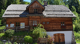 Ein traditionelles Holzhaus mit einem steilen Dach und Fensterläden. Umgeben von üppigem Grün und Pflanzen.