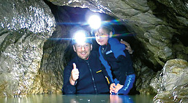 Ein Mann und ein Kind stehen in einer Höhle und tragen Stirnlampen. Sie stehen im Wasser und lächeln, während der Mann mit dem Daumen nach oben zeigt.